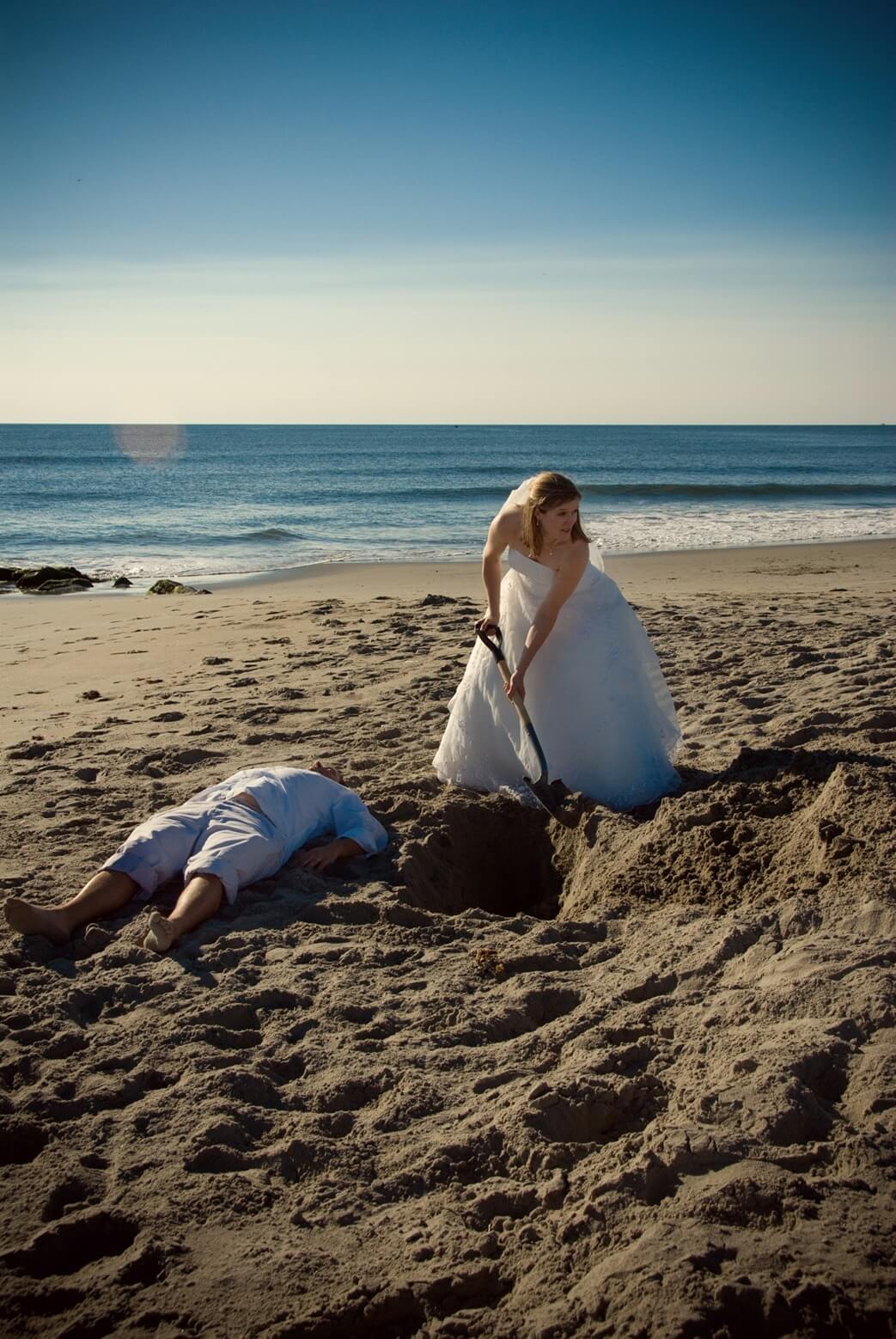 ri-trash-the-dress-photographer-005 A woman in a wedding dress is digging a hole in the sand on a beach, while a man lies face down next to the hole.