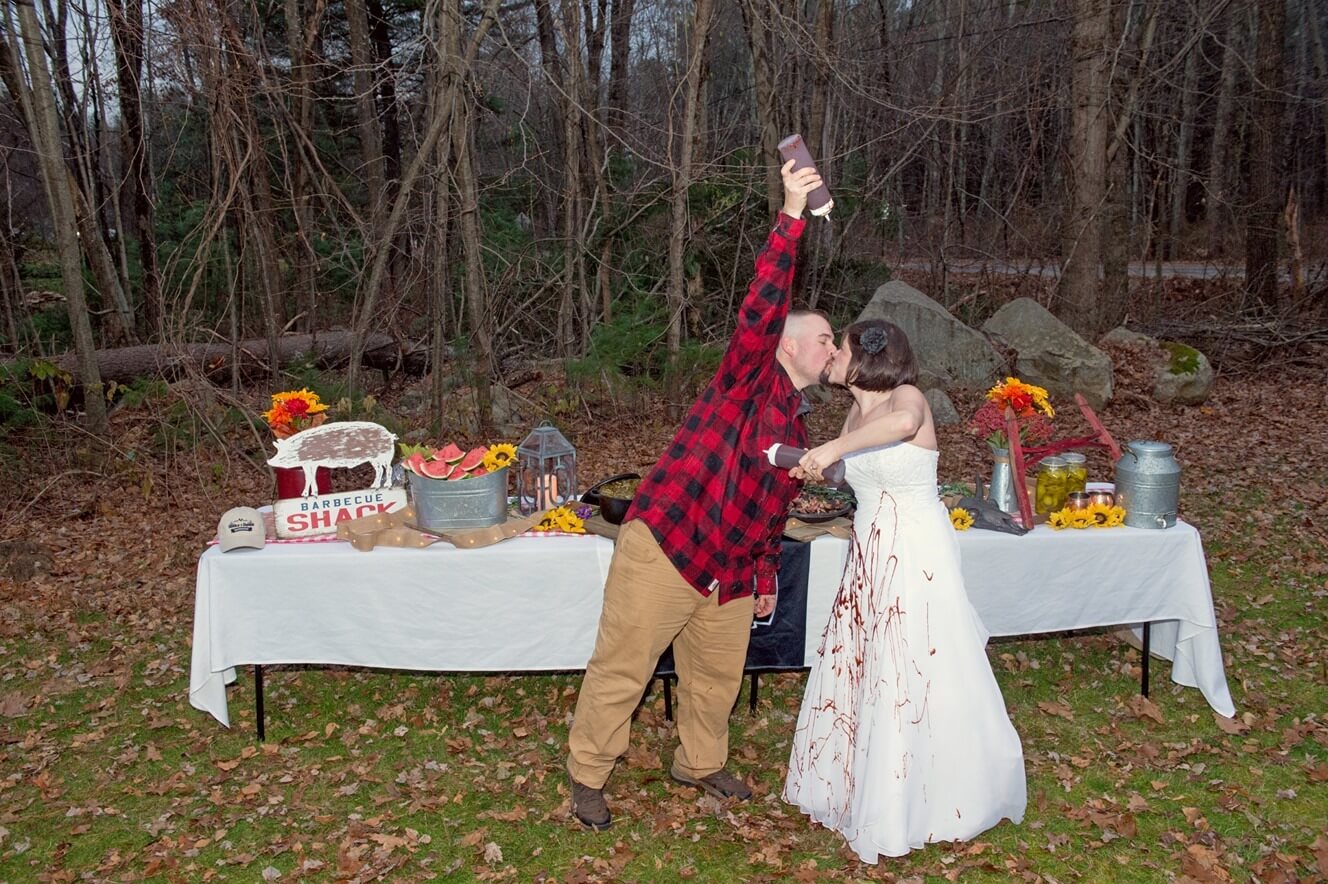 ri-trash-the-dress-photographer-006 A man in a plaid shirt and a woman in a white dress kiss in front of an outdoor table with food and fall decorations; the man holds a bottle aloft.