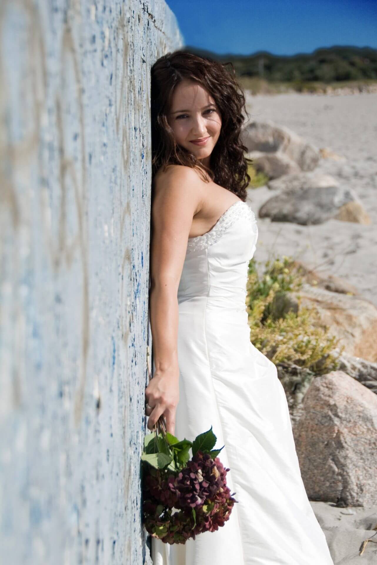 ri-trash-the-dress-photographer-007 A woman in a white wedding dress leans against a weathered wall on a beach, holding a bouquet of dark flowers at her side.