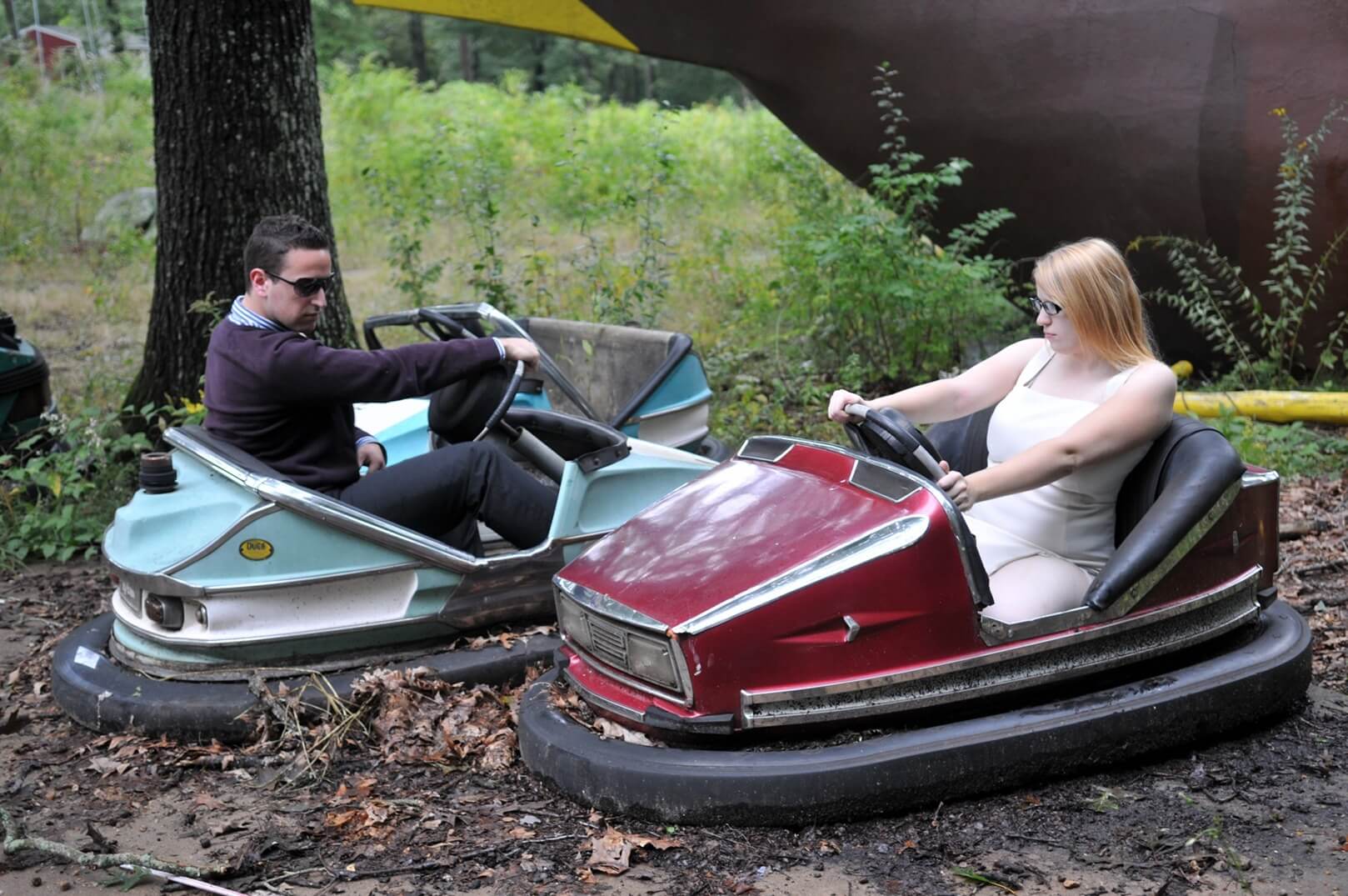 ri-trash-the-dress-photographer-008 Two adults sit in stationary bumper cars outdoors in a wooded area, surrounded by fallen leaves and overgrown vegetation.