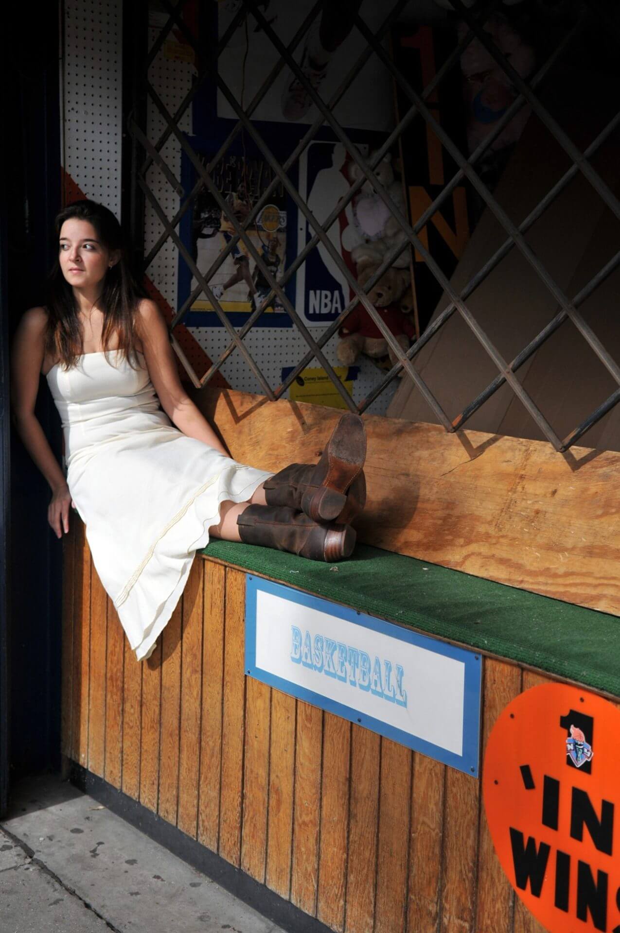 ri-trash-the-dress-photographer-009 A woman in a white dress and brown boots sits on a counter with her legs stretched out, next to a sign that reads "BASKETBALL" at an outdoor booth.