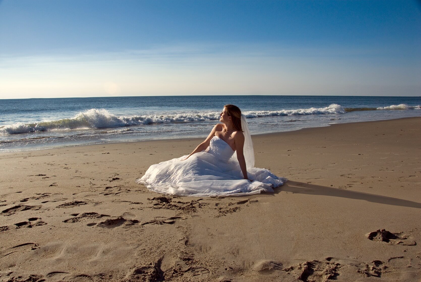 ri-trash-the-dress-photographer-010 A woman in a white wedding dress sits on the sandy shore of a beach, looking towards the ocean under a clear blue sky.