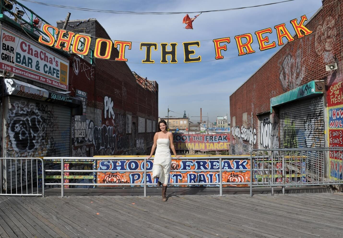 ri-trash-the-dress-photographer-011 A woman in a white dress stands on a wooden boardwalk under a "SHOOT THE FREAK" sign, surrounded by graffiti-covered buildings and closed storefronts.