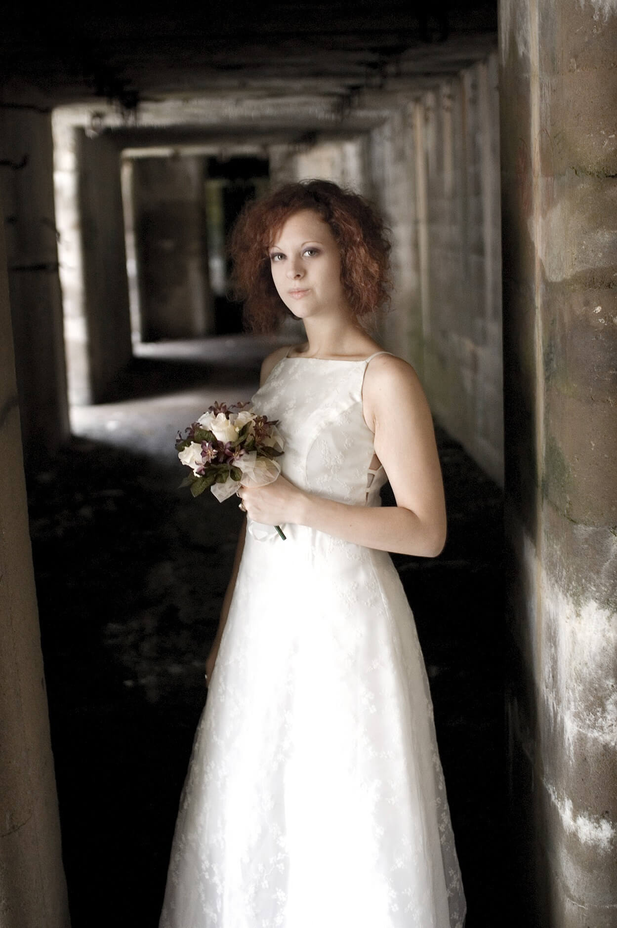 ri-trash-the-dress-photographer-012 A woman in a white sleeveless wedding dress holds a small bouquet of flowers while standing in a dimly lit corridor.