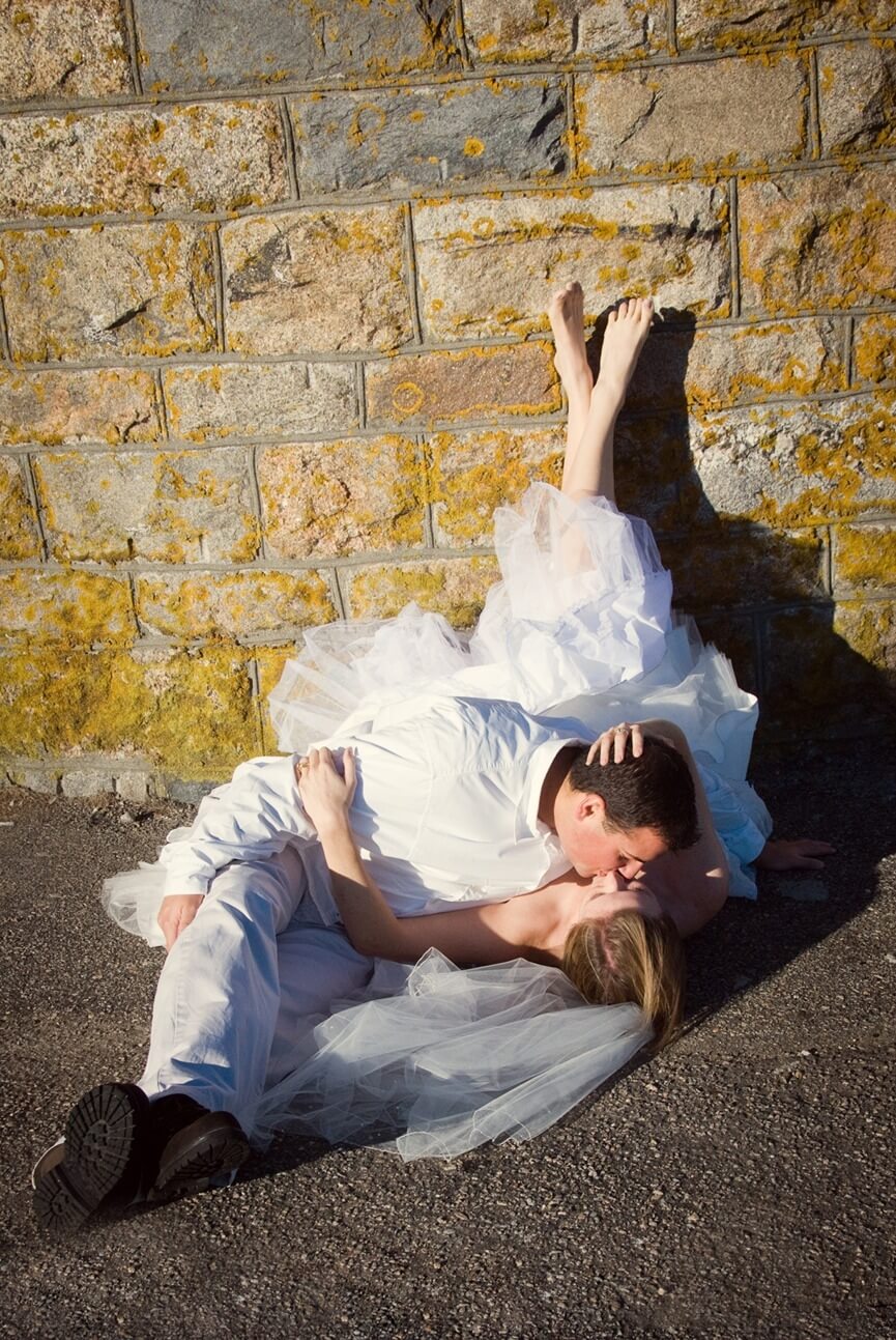 ri-trash-the-dress-photographer-013 A bride and groom in wedding attire lie on the ground by a stone wall, kissing, with the bride's legs raised and leaning against the wall.