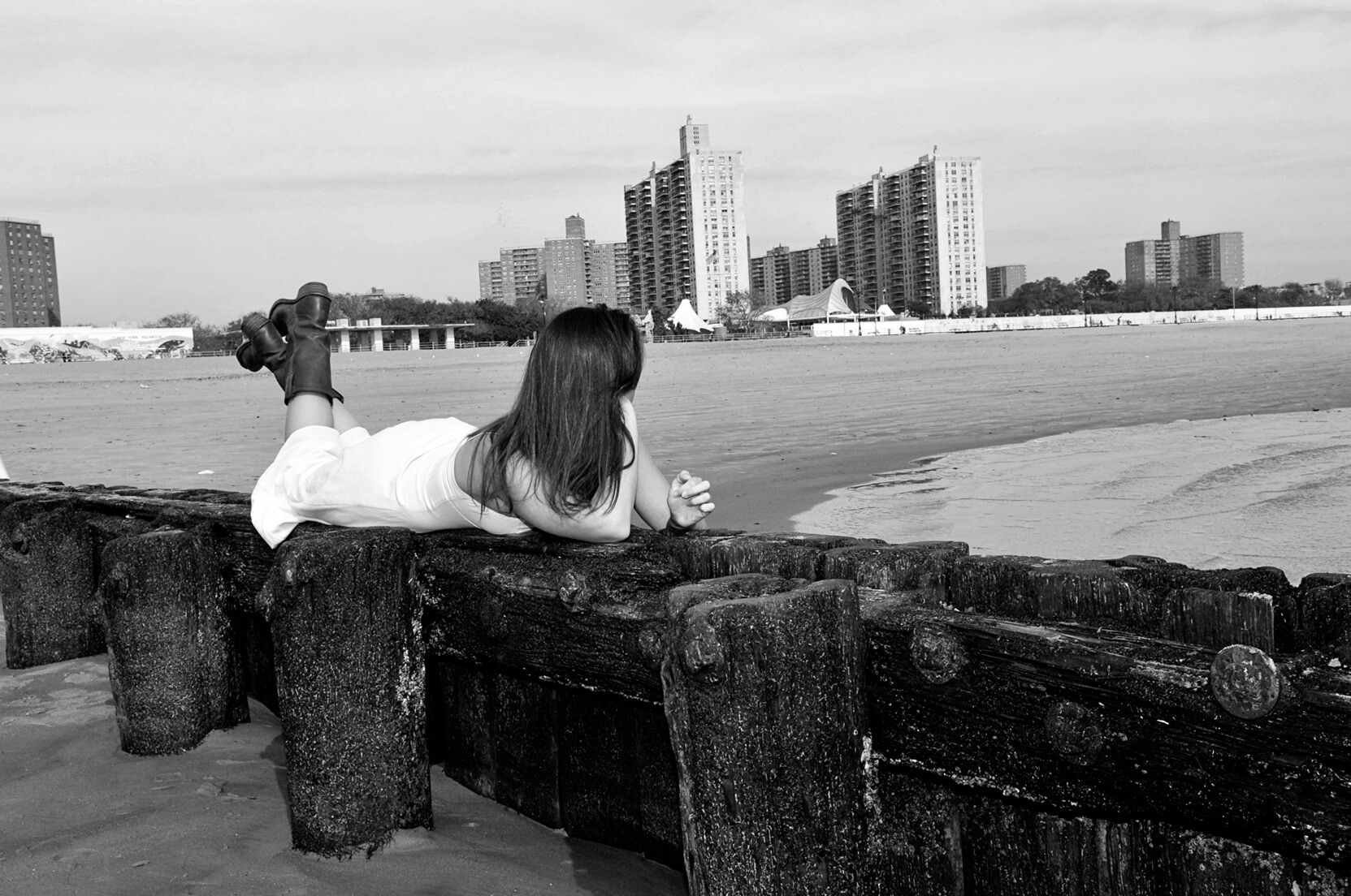 ri-trash-the-dress-photographer-014 A woman in a white dress lies on a wooden pier facing the ocean, with city buildings visible in the background. The image is in black and white.