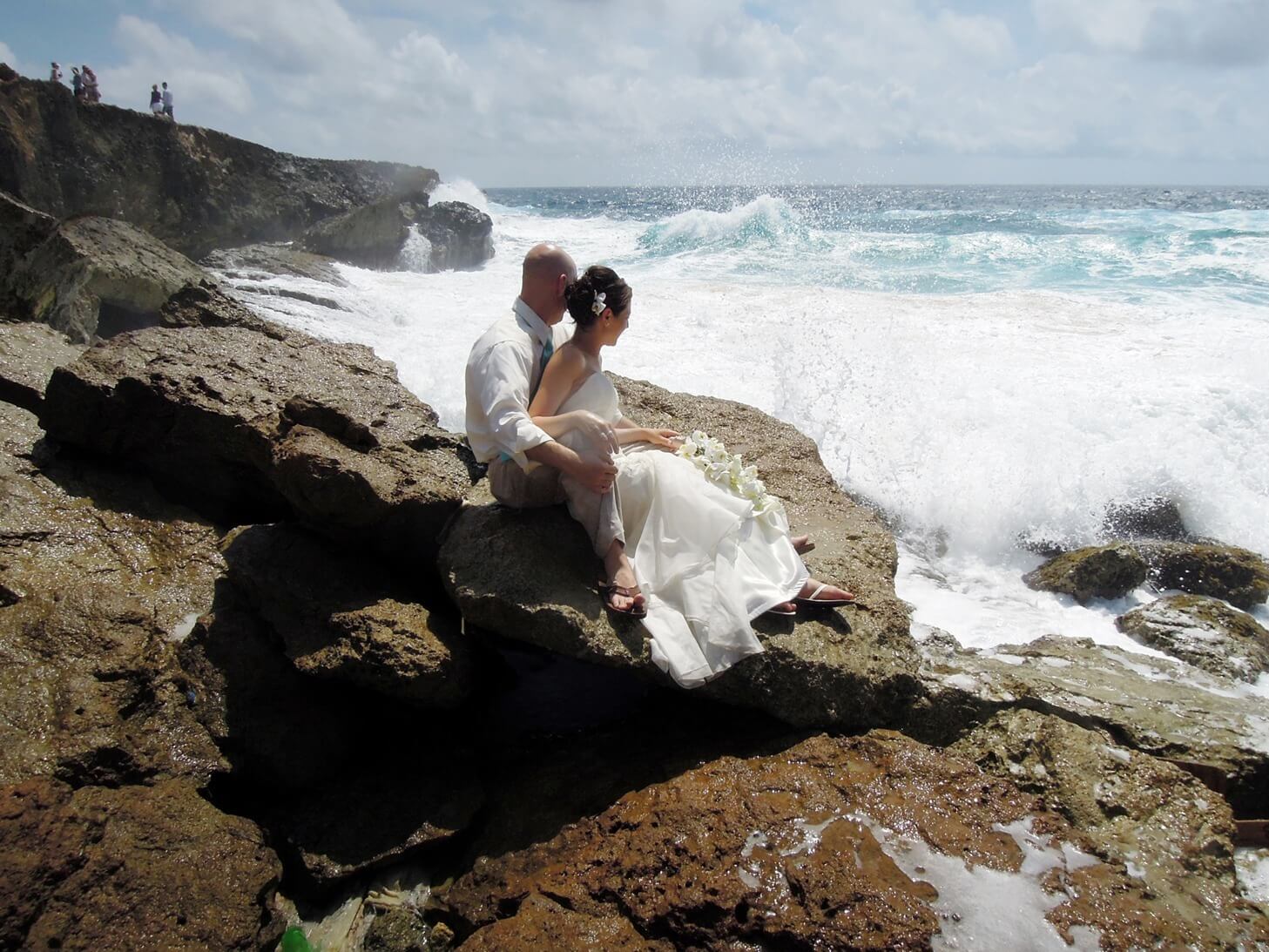 ri-trash-the-dress-photographer-015 A bride and groom sit on large rocks by the ocean with waves crashing nearby, both facing the sea under a partly cloudy sky.
