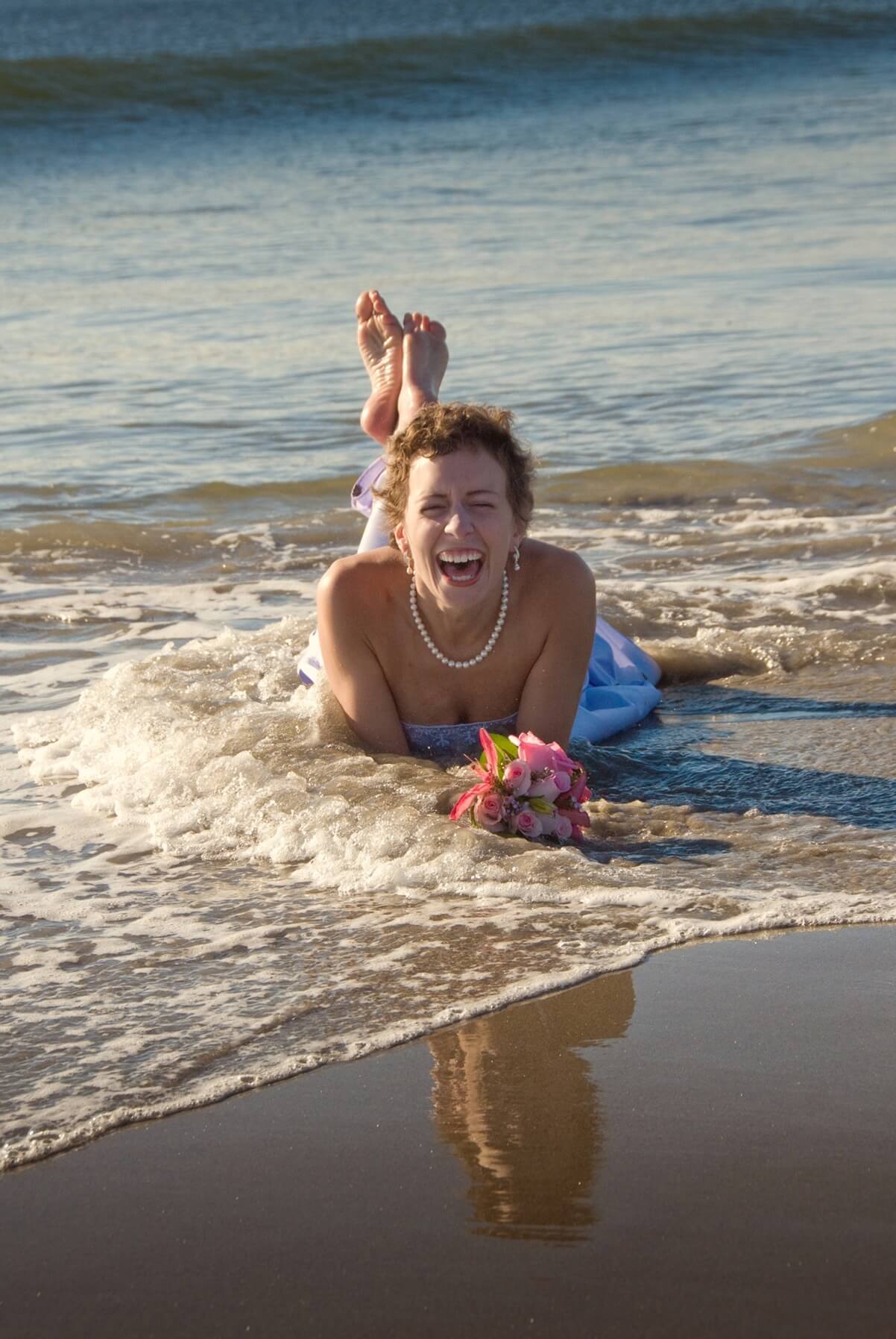 ri-trash-the-dress-photographer-017 A woman in a white dress and pearl necklace lies on her stomach in shallow ocean water, smiling and holding a pink bouquet.
