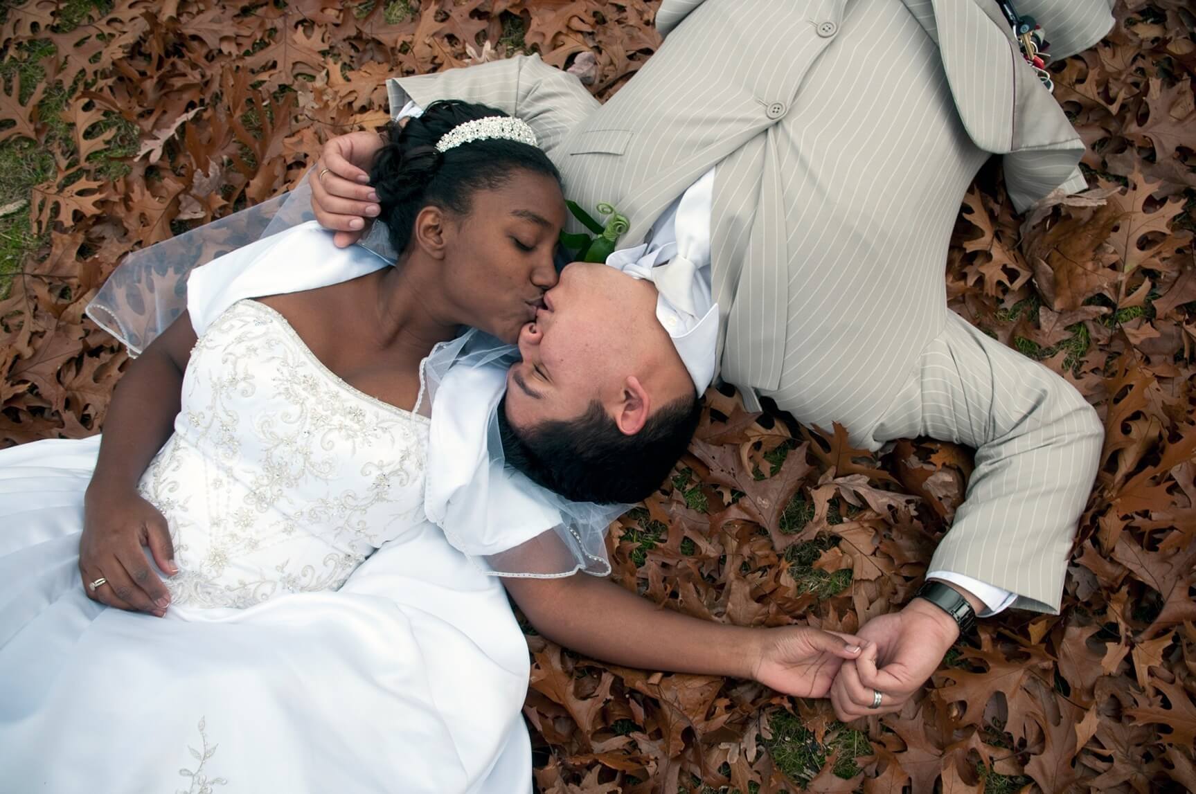 ri-trash-the-dress-photographer-018 A bride and groom lie on fallen leaves, holding hands and kissing, dressed in wedding attire.