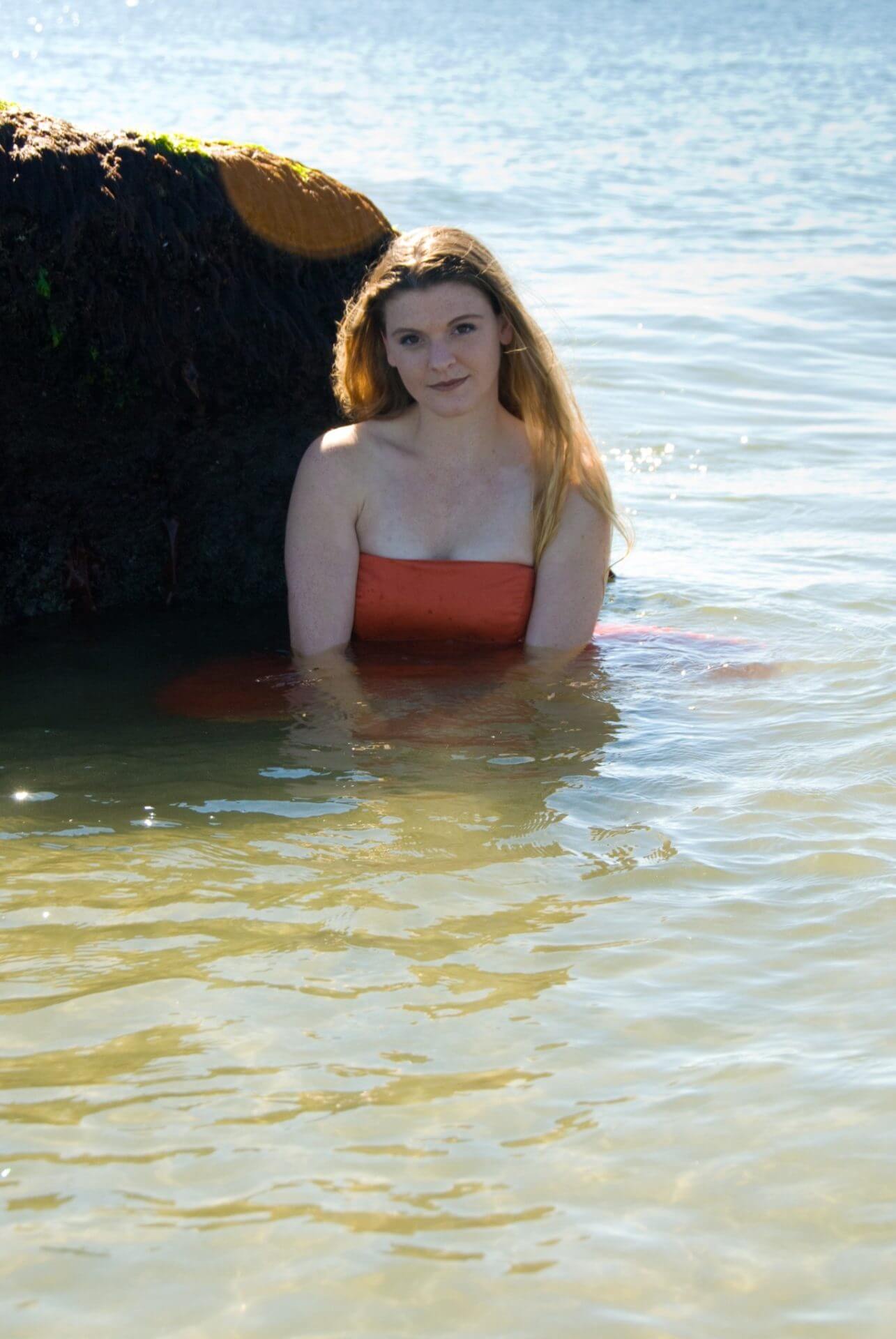 ri-trash-the-dress-photographer-019 A woman in a red strapless top and long hair sits partially submerged in shallow water near a large rock at the beach.