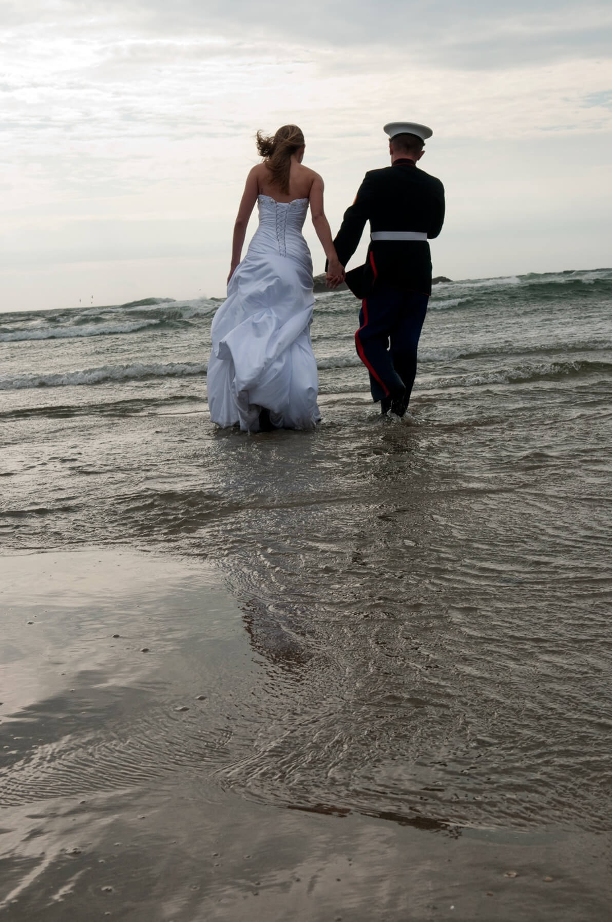ri-trash-the-dress-photographer-020 A bride in a white dress and a person in a military uniform walk hand in hand toward the ocean on a cloudy day.