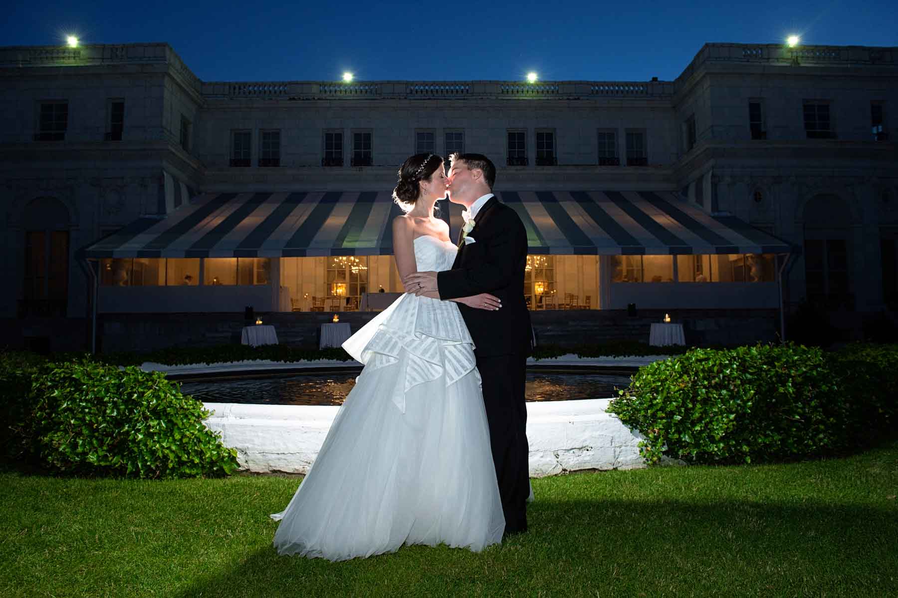 A bride and groom stand together in front of a lit building at night, embracing and gazing at each other on the lawn.