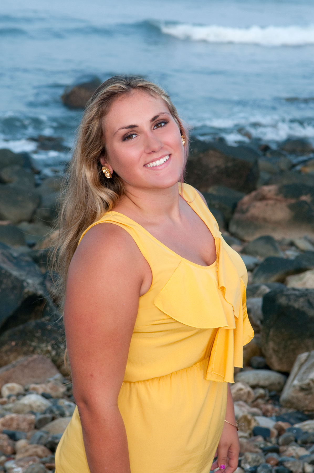 senior-portrait-girl-in-yellow-by-rocks-at-ocean A woman in a yellow dress stands on a rocky beach, smiling at the camera with waves and rocks in the background.