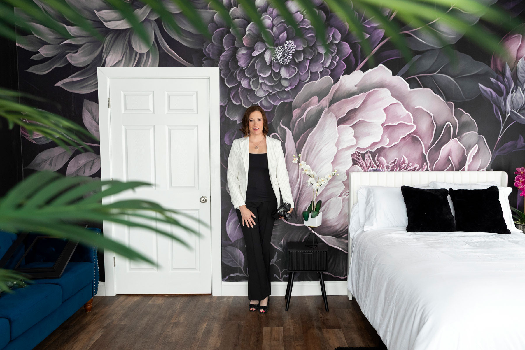 warwick-photography-studio-nicole-in-boudoir Woman in a white blazer stands next to a bed in a modern bedroom with large floral wall art, wooden floor, and plants in the foreground.